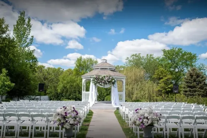 Outdoor wedding ceremony setup with white chairs, a gazebo, and flower arrangements. Bright blue sky.