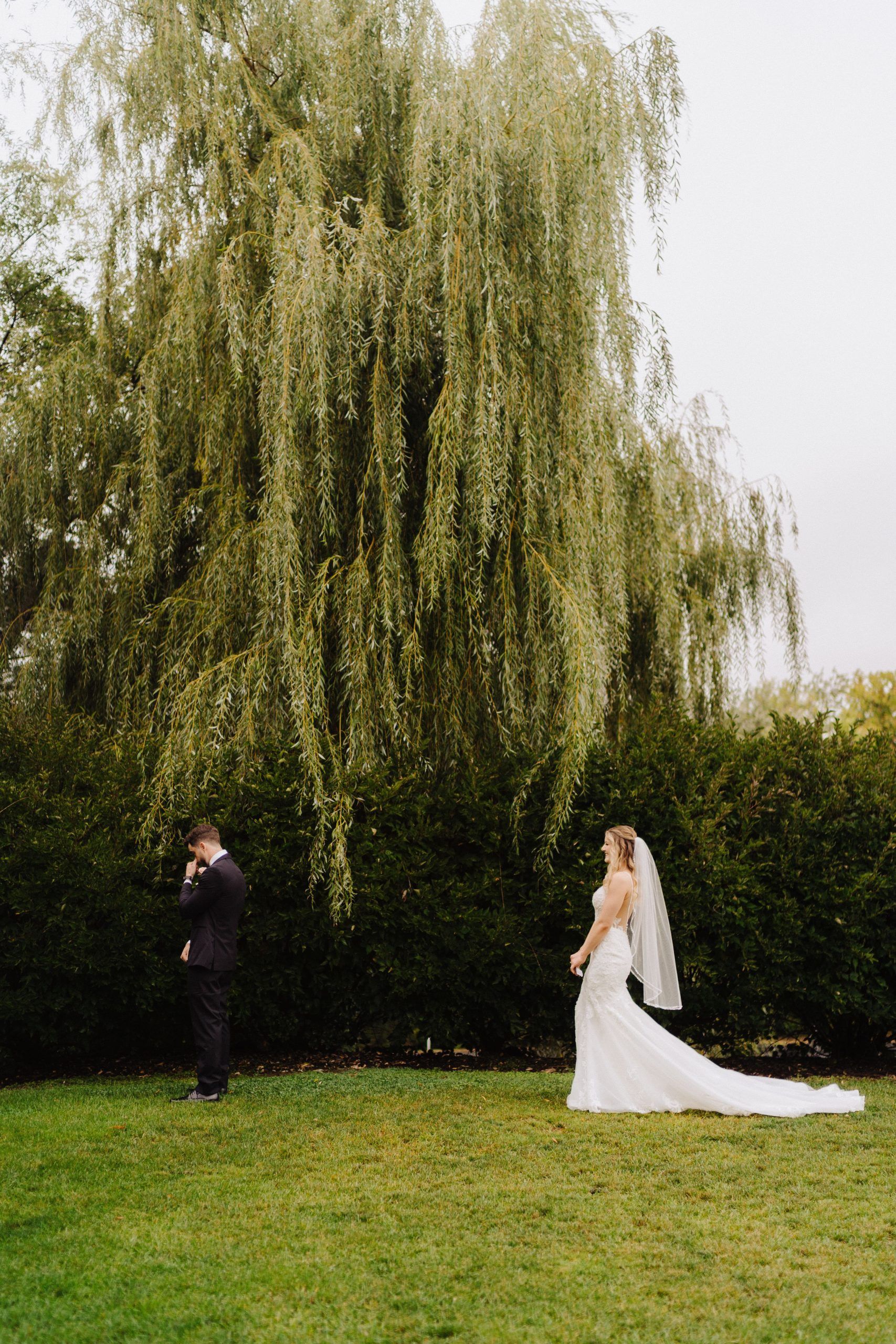 Bride in white gown facing groom under a weeping willow tree. Groom in a dark suit covering his face.