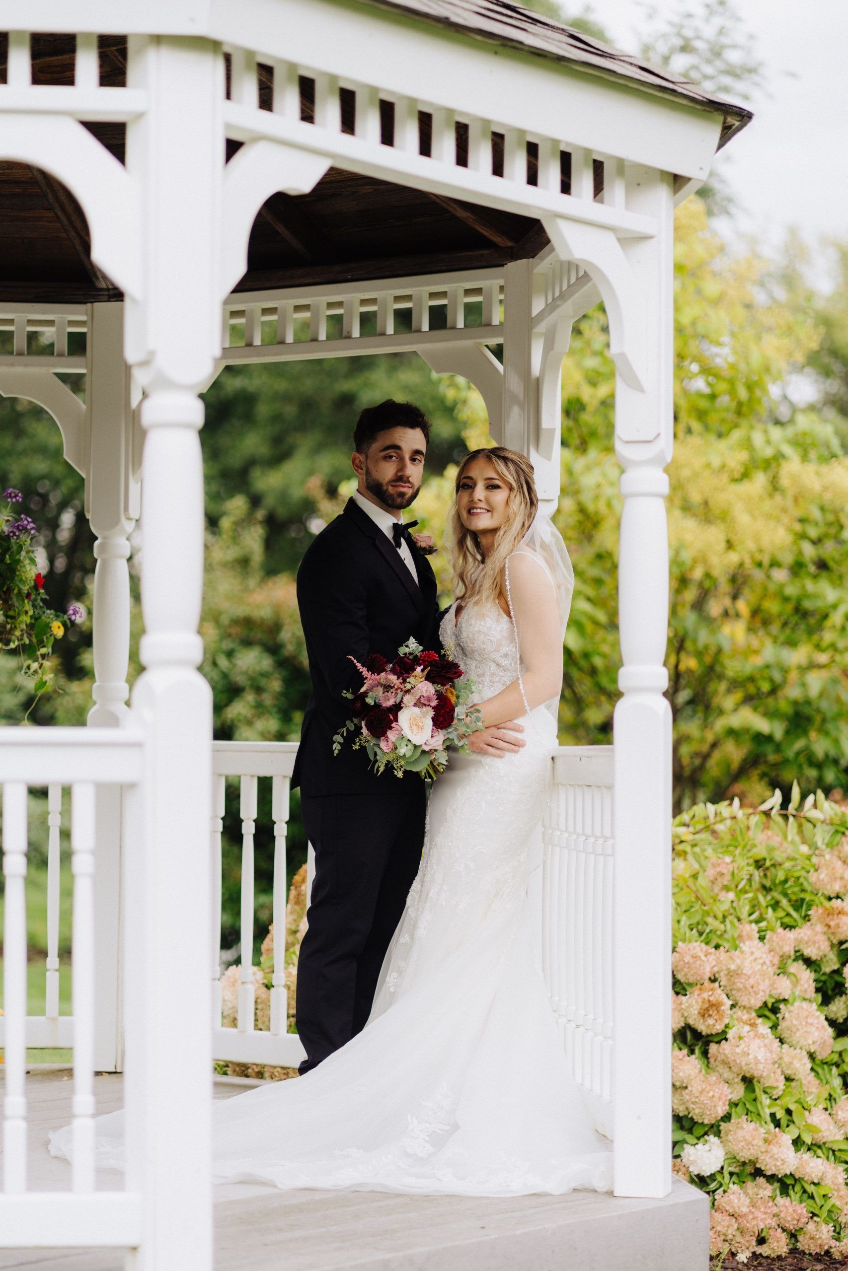 Bride and groom pose in a white gazebo. The bride holds flowers. The groom wears a suit. Green trees in background.