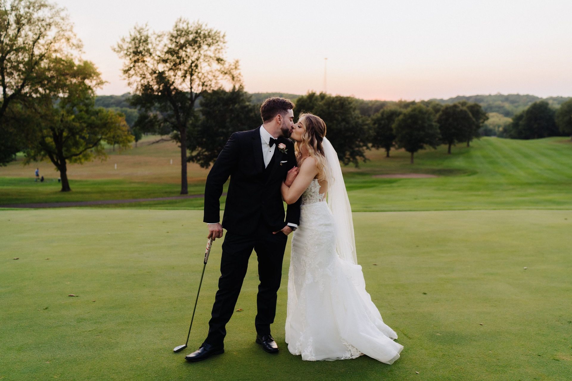 Newlyweds on golf course: groom kissing bride's temple, holding golf club; green grass, trees, sunset backdrop.
