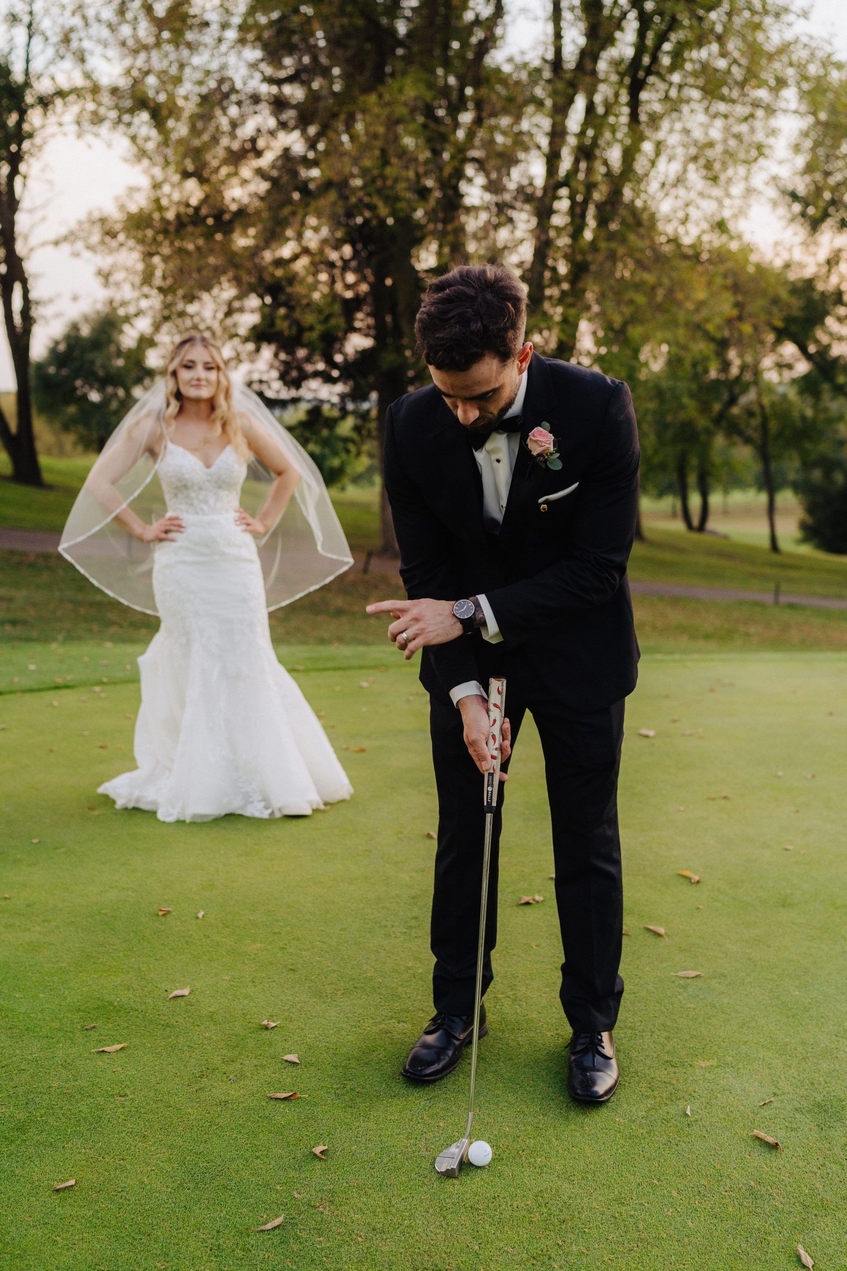 Groom in a tuxedo putts golf ball as bride watches from the background on a golf course.