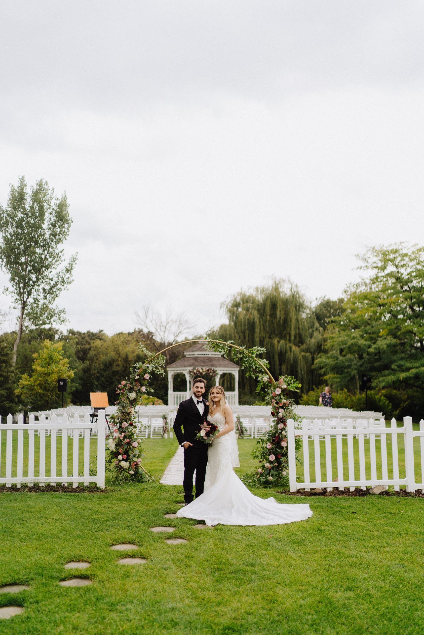 Bride and groom pose near a white picket fence at their outdoor wedding.