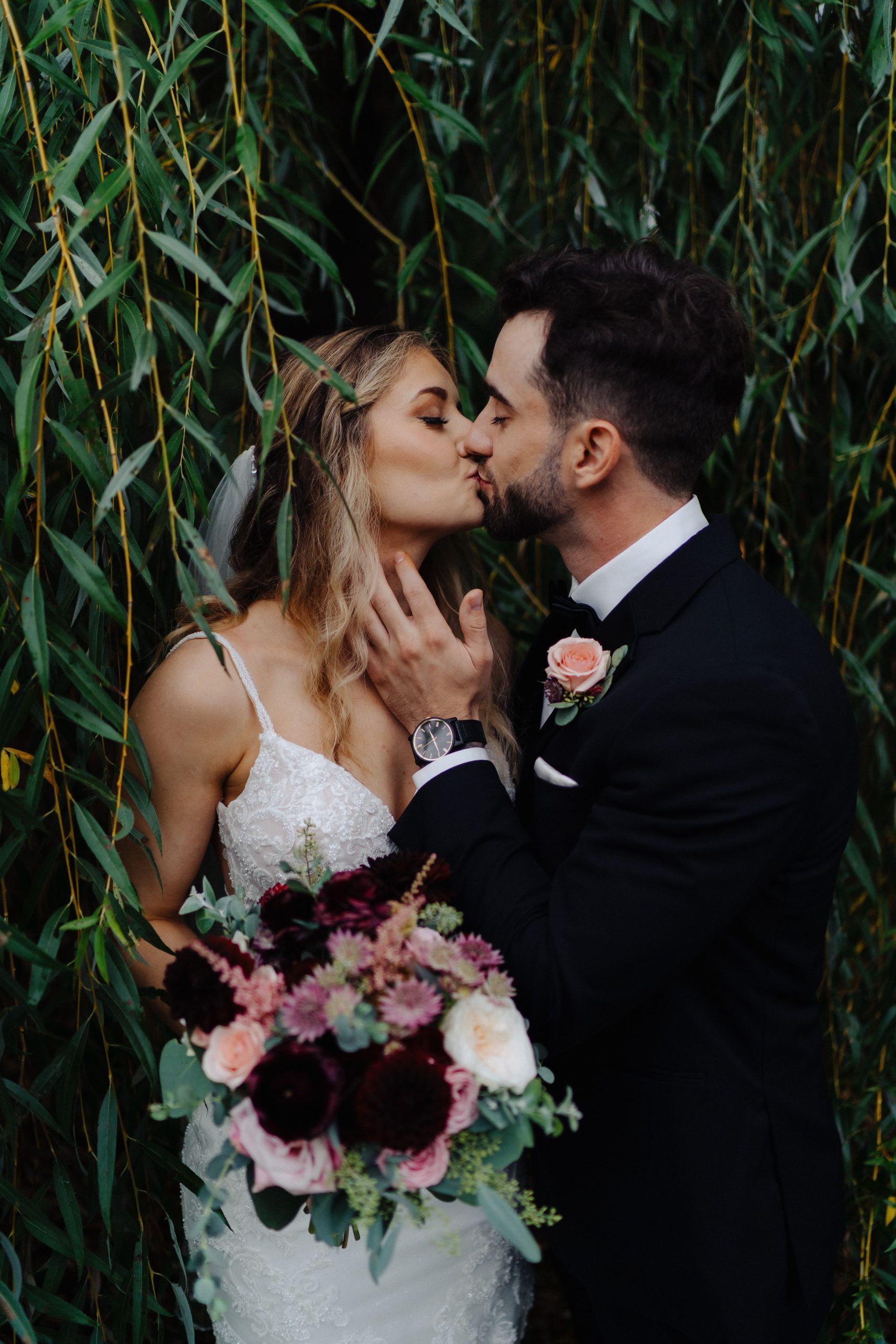Bride and groom kissing under weeping willow. Bride in white dress, holding bouquet; groom in black suit.