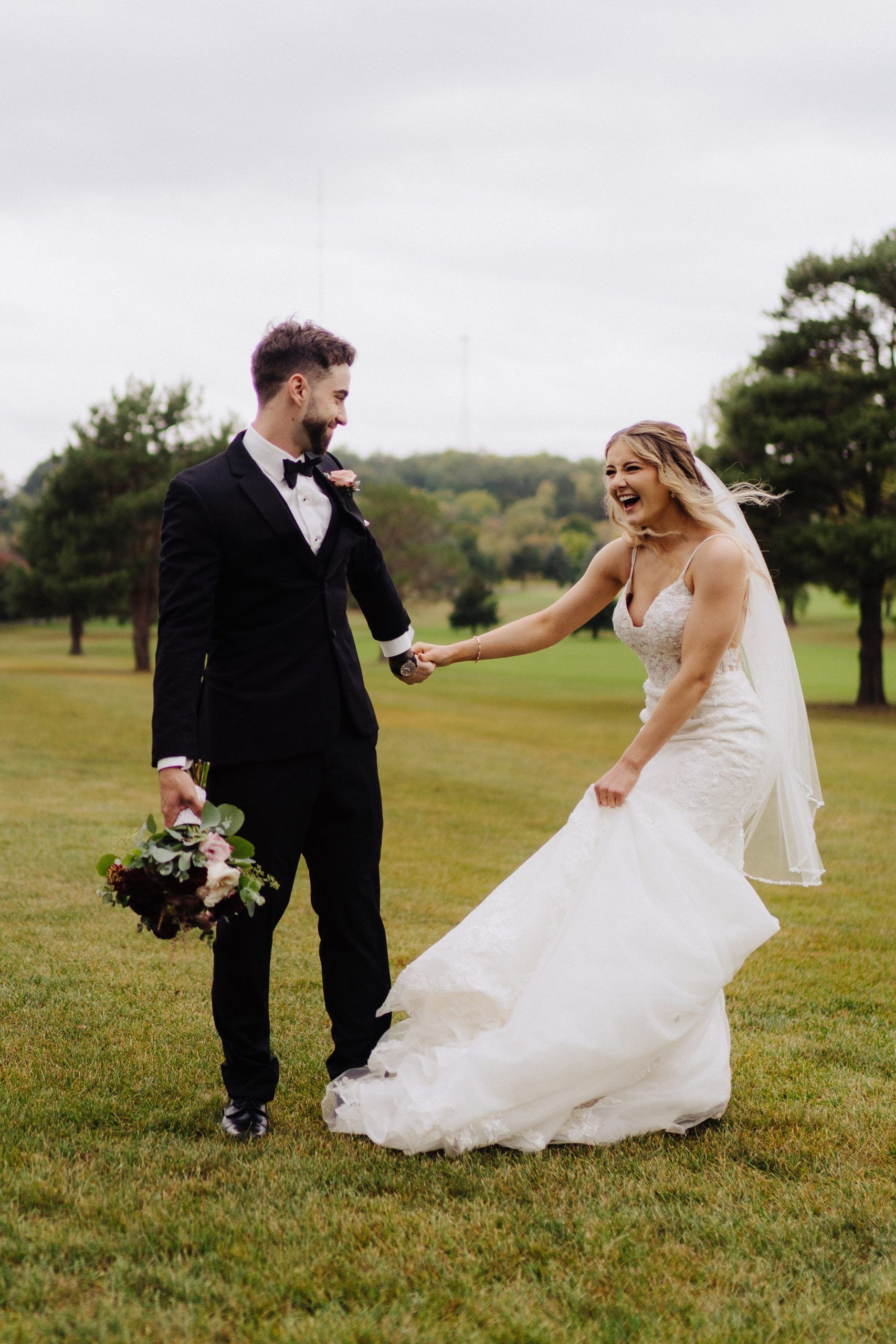 Bride and groom holding hands, laughing on a green field. Bride in a white dress, groom in a black suit.