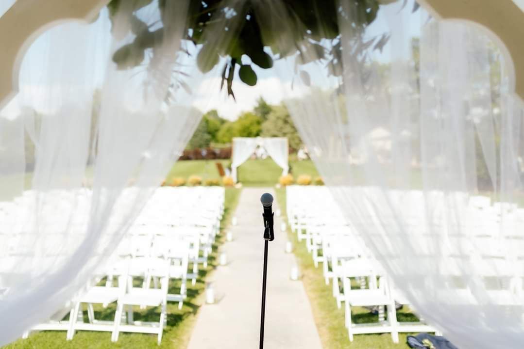 Outdoor wedding ceremony setup with white chairs, arch, microphone, and greenery.