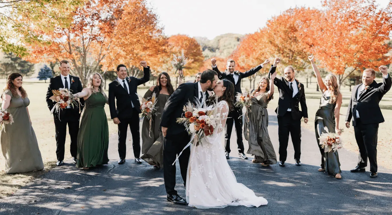 Wedding party celebrating with a kiss in front, fall foliage in the background, with wedding party wearing dark formal wear, embracing.