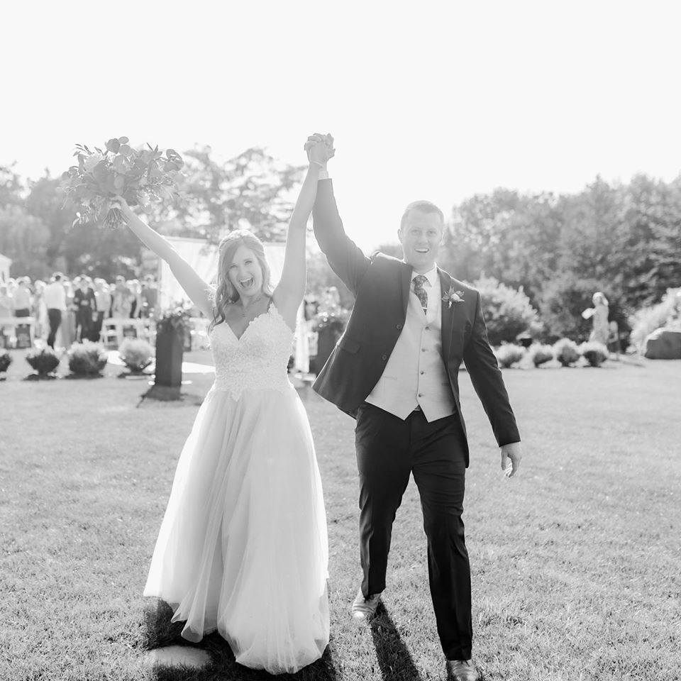 Newlyweds raise arms celebrating, walking outdoors. Bride in white dress, groom in suit, sunny garden setting.