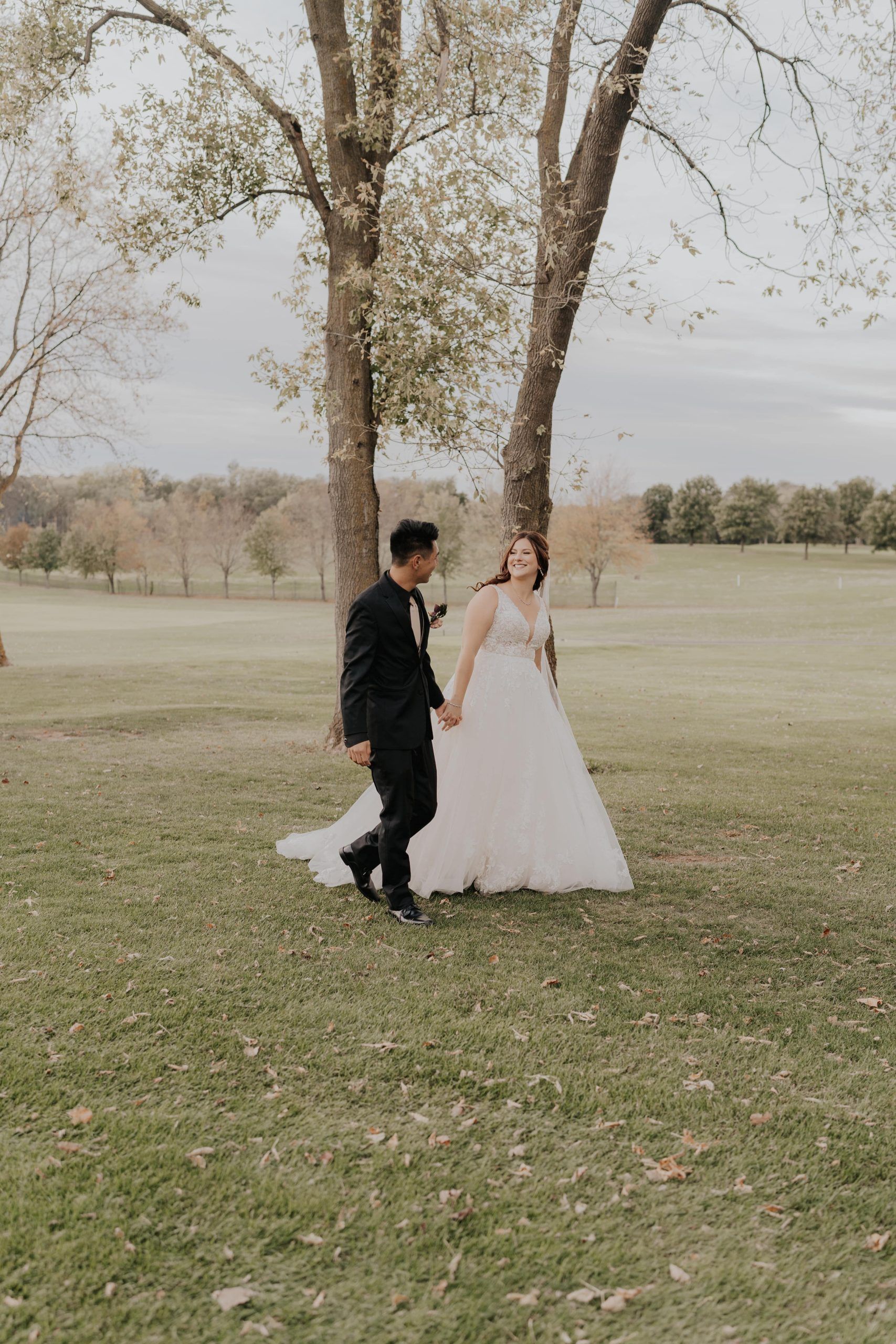 A newly married couple holds hands walking through a grassy field. The bride smiles, wearing a white gown. The groom wears a black suit.