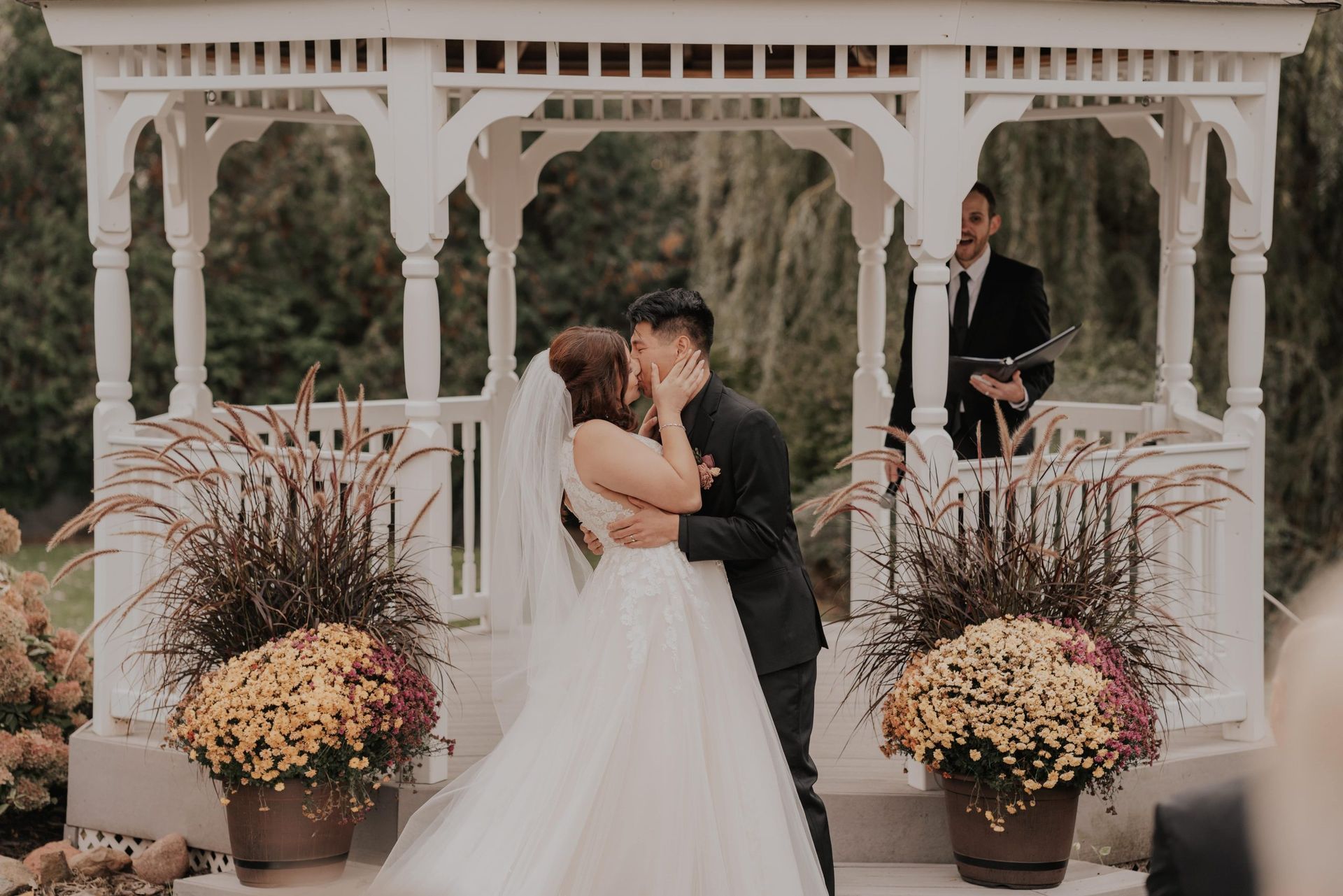Couple kissing at a wedding ceremony in a white gazebo. A guitarist plays in the background.
