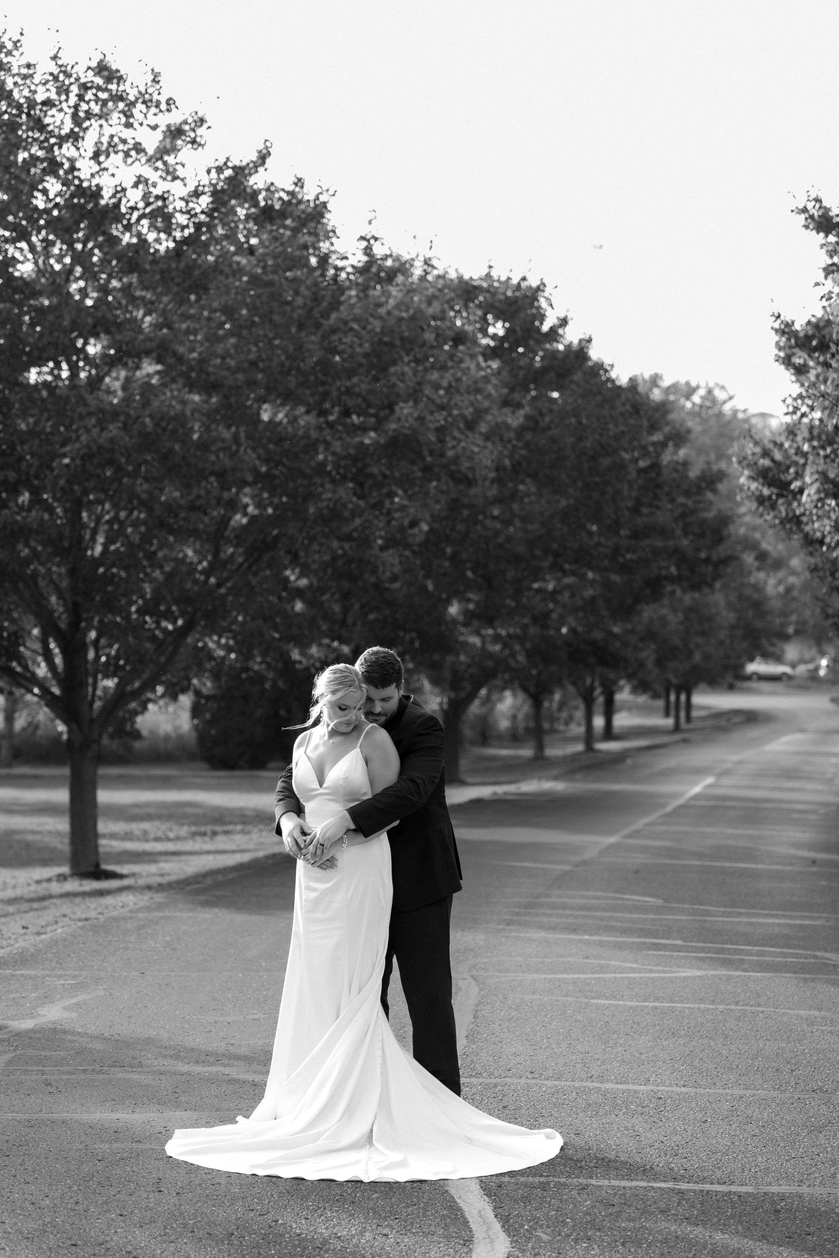 Bride and groom embrace on a road lined with trees; black and white.