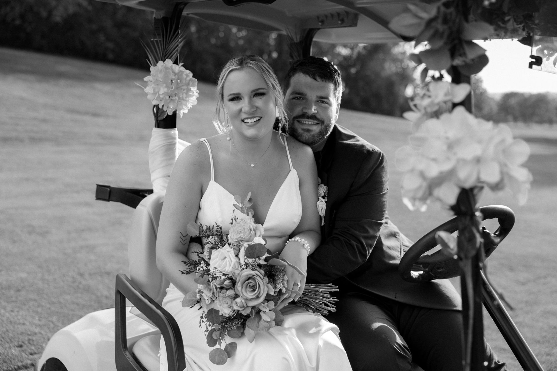 Newlyweds in decorated golf cart, bride holding bouquet smiles, groom smiles, outdoors.
