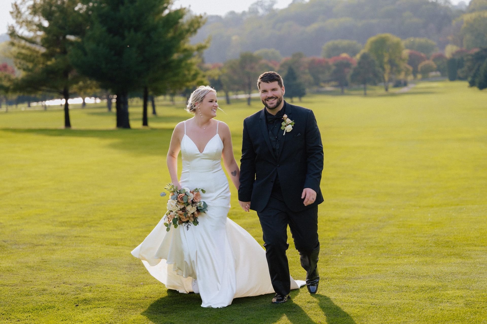 Newlyweds walking on a grassy lawn, holding hands. Blonde bride in white dress, groom in black suit, trees in background.