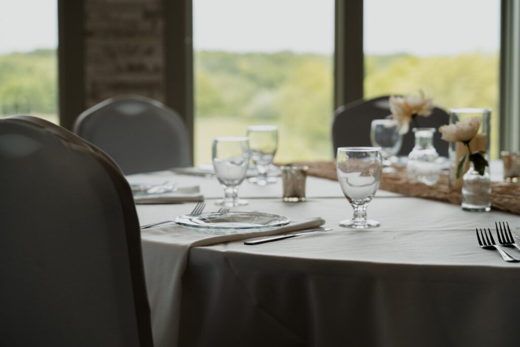 Dining table set for a celebration with glasses, silverware, and flowers, with a view of nature in the background.