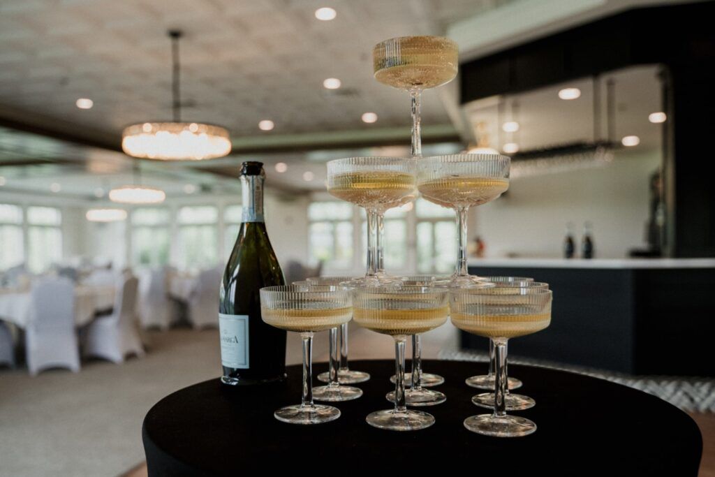 Champagne tower with bottle, set on a black table in a banquet room, with white tablecloths and a bar in the background.