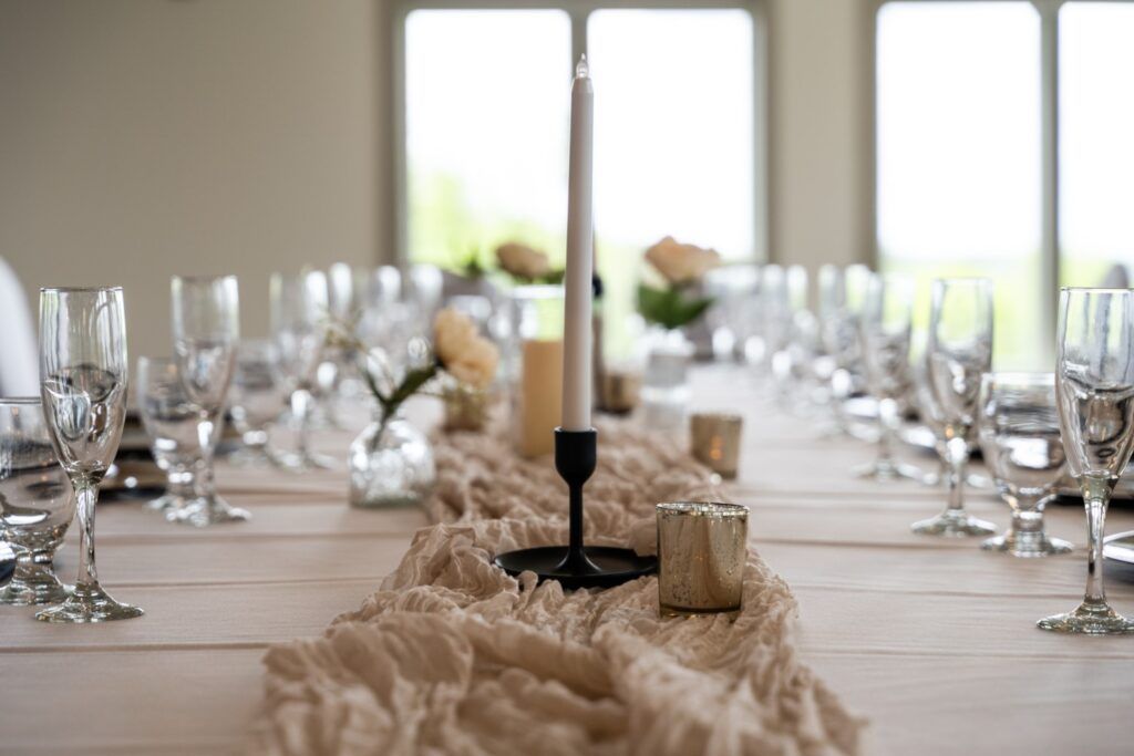 Long table set for a dinner party with glassware, a candle centerpiece, and a neutral-colored runner.