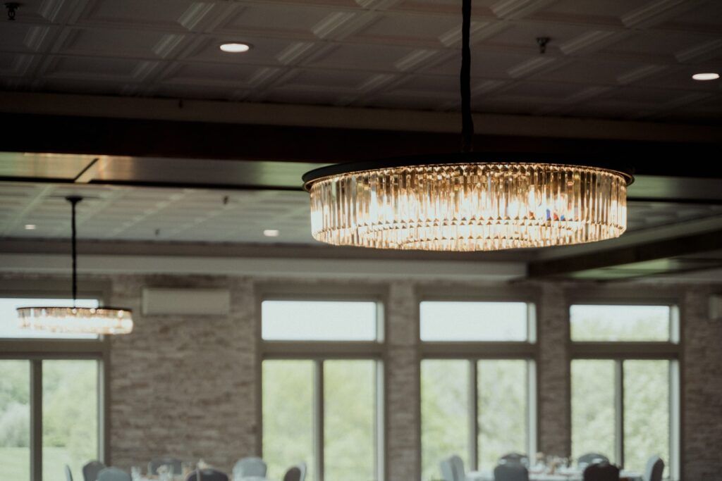 Elegant chandelier hanging in a room with large windows and brick walls, near tables and chairs.