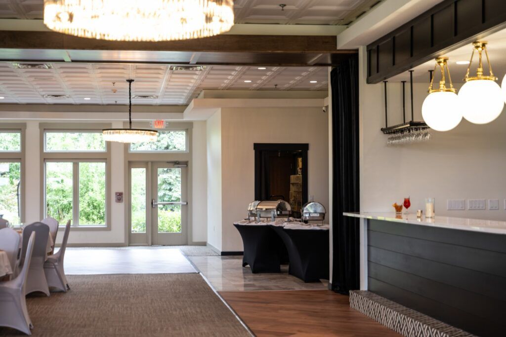 Elegant event space with a bar, tables, and large windows. Neutral colors, chandeliers, and a wooden floor.
