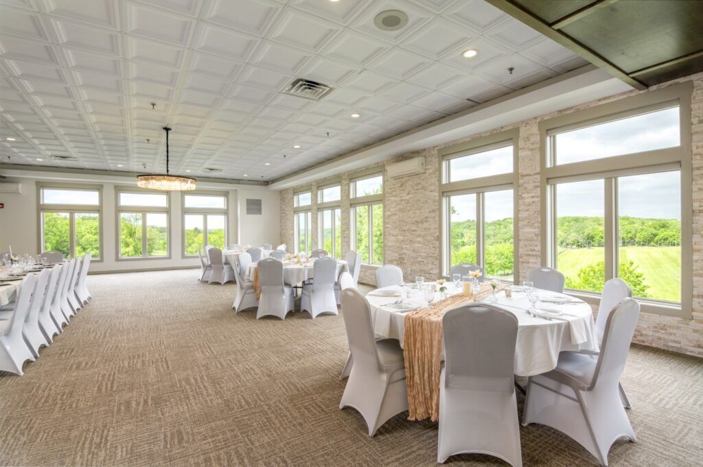 Banquet hall with round tables set for a formal event; windows overlook a green field.
