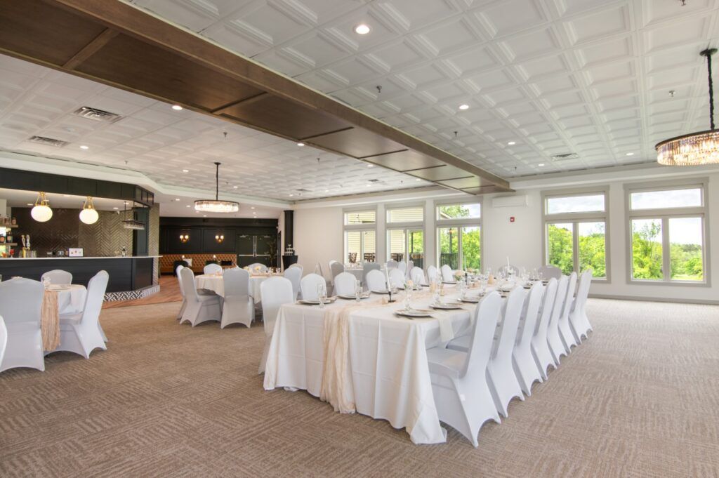 A large banquet room with tables set for a formal event. Bright, neutral colors and windows.