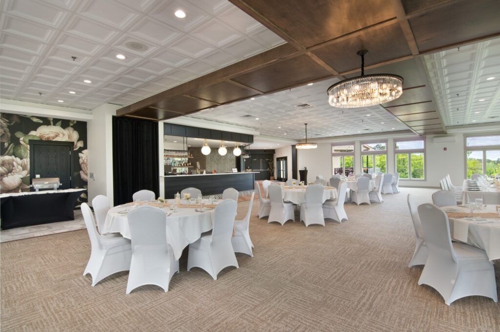 Banquet hall with round tables set for guests, white chair covers, beige carpet, floral wall art, and a bar in the background.