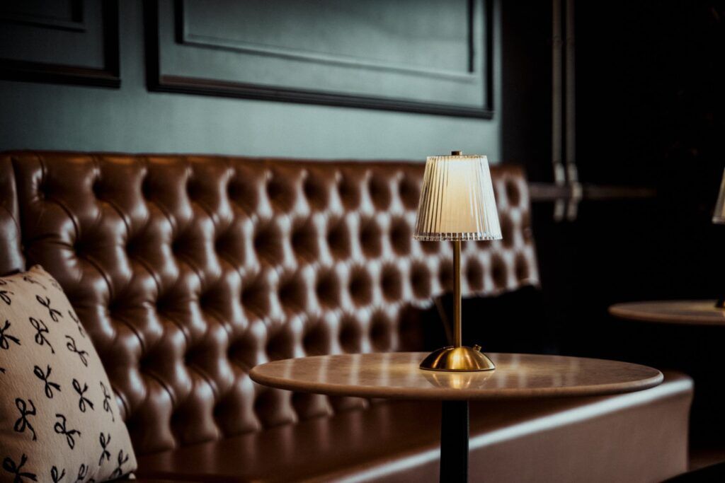 Cozy booth in a dimly lit restaurant. Brown leather tufted bench, small round table with a lamp.