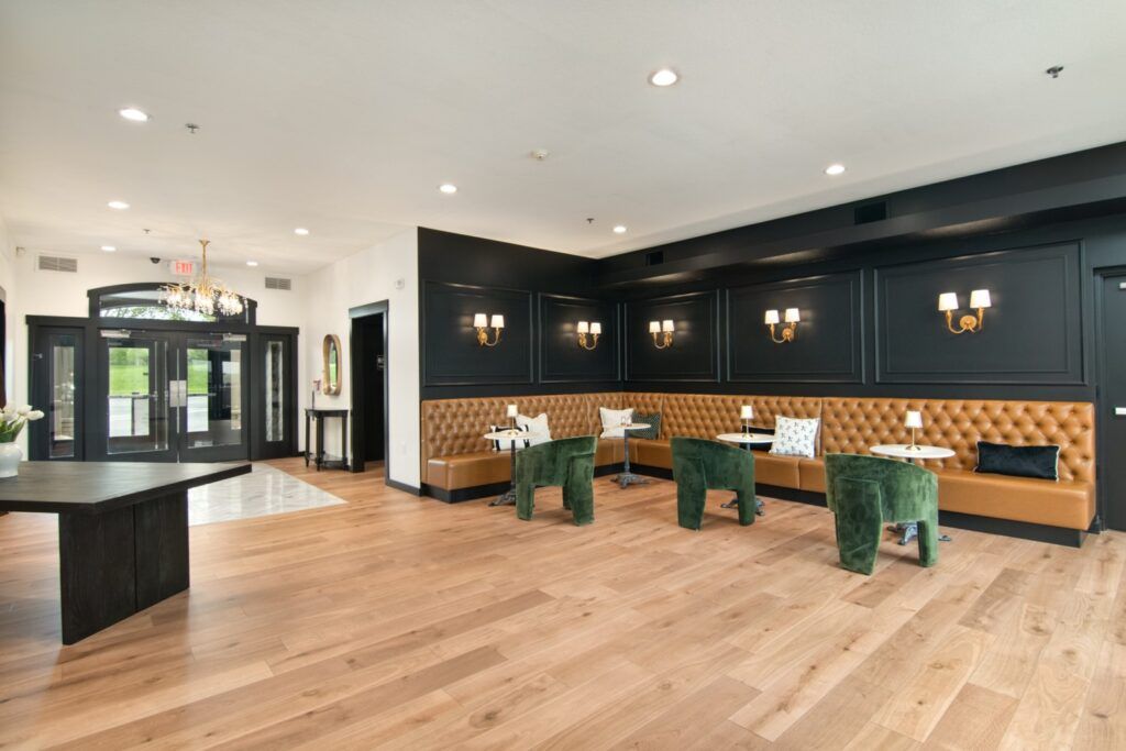 Lounge area with wood floors, dark accent wall, tan booth seating, and green velvet chairs. Glass doors in background.