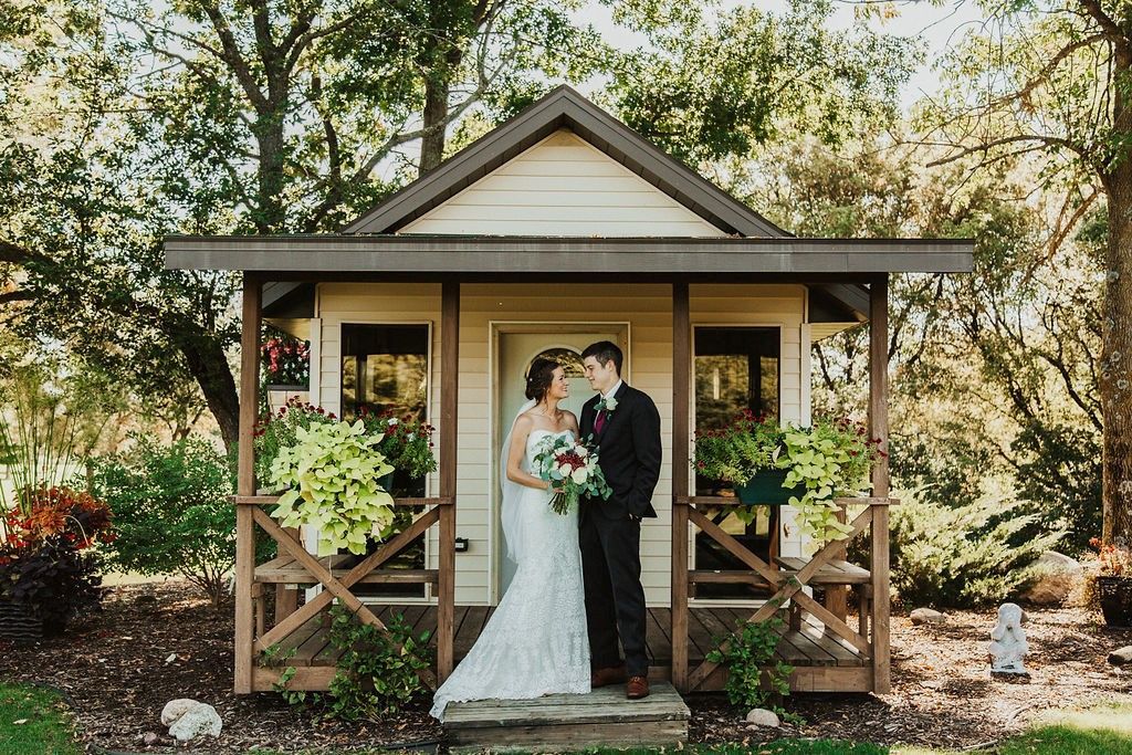 Bride and groom stand at the door of a small, wood-framed house, surrounded by greenery.