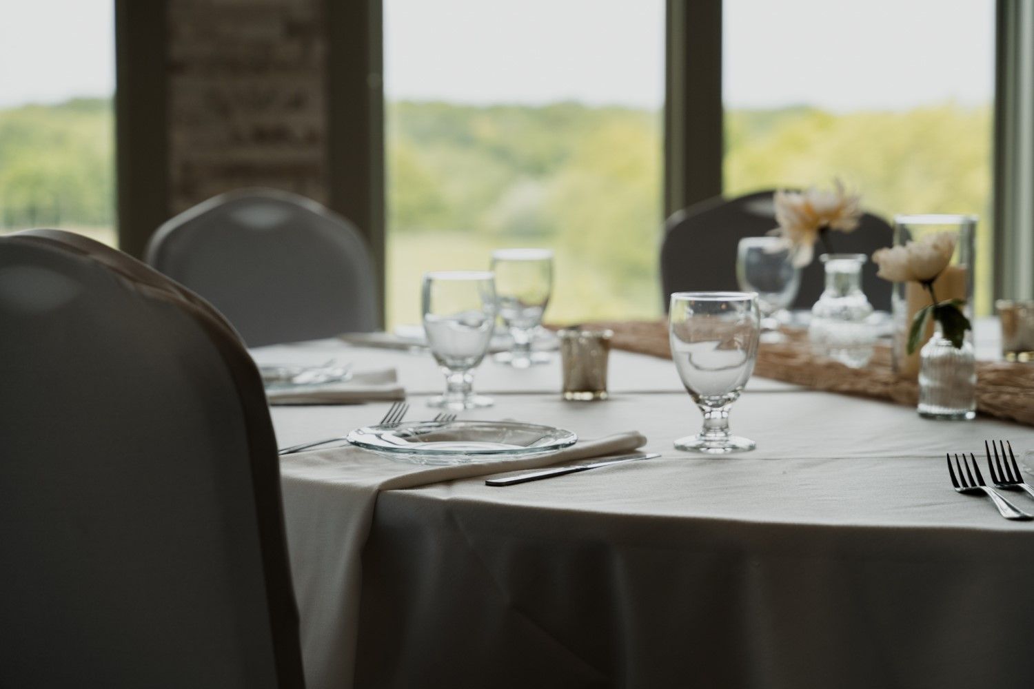Round table set for a formal event, with white tablecloth, glassware, and floral arrangement; view of green trees in the background.