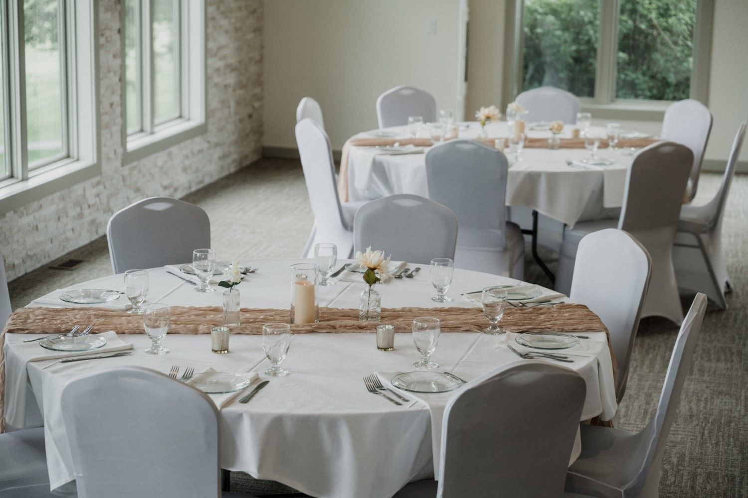 Round tables set for a meal in a light-filled room with white chair covers.