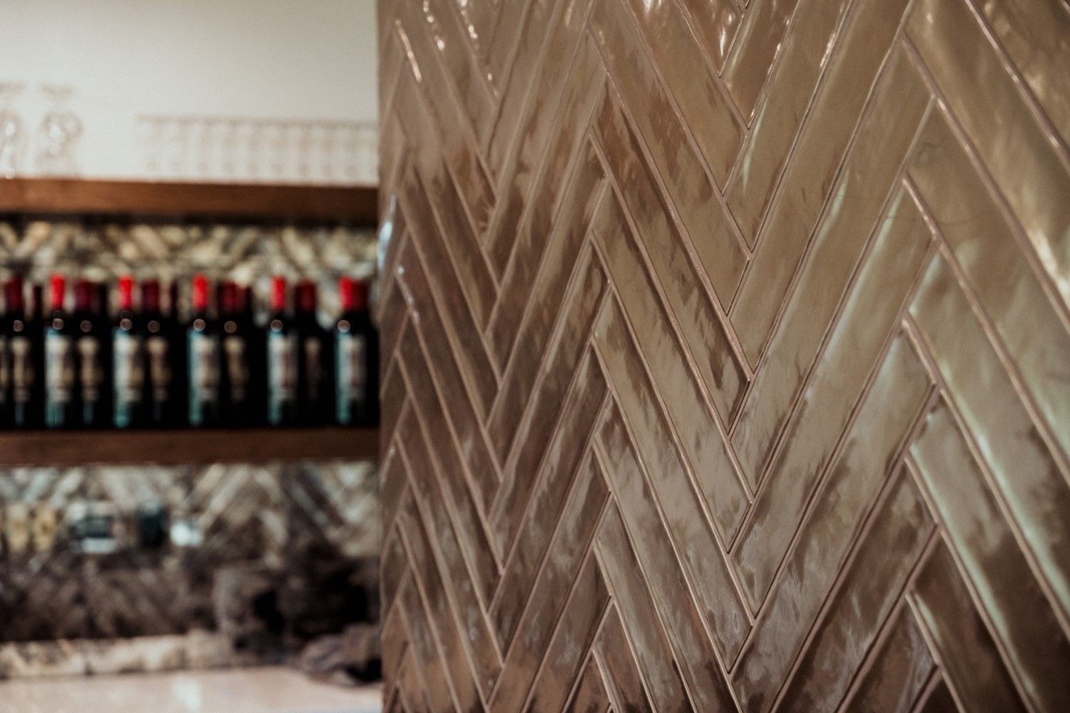 Herringbone-patterned brown tile wall with blurred background of wine bottles and shelves.