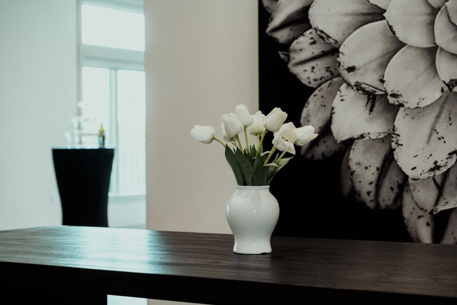 White tulips in a white vase on a dark wood table; black and white artwork in the background.