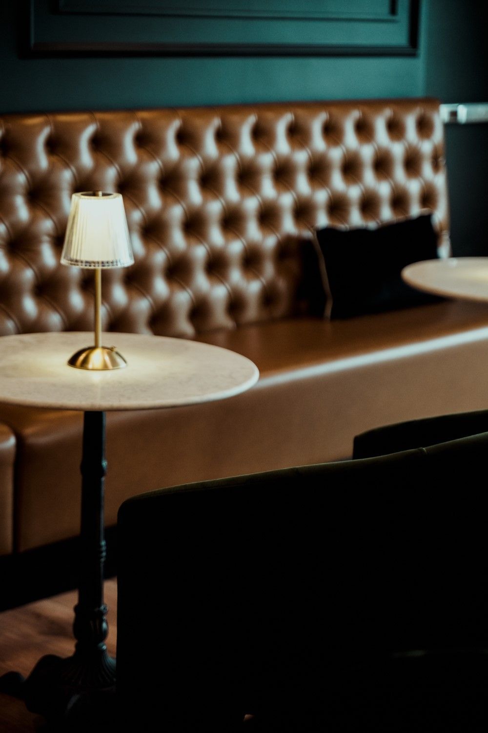 A restaurant booth with a marble table, small lamp, and brown tufted leather back.