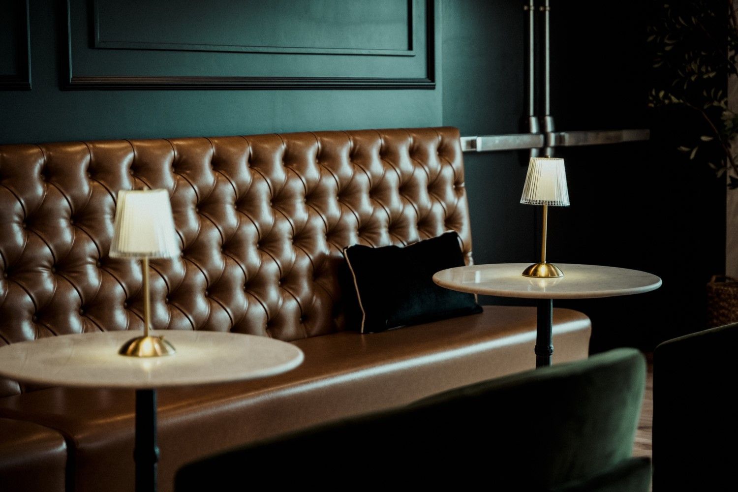 Cozy bar booth with tufted brown leather bench, marble tables, and small lamps, green walls and a dark, intimate atmosphere.