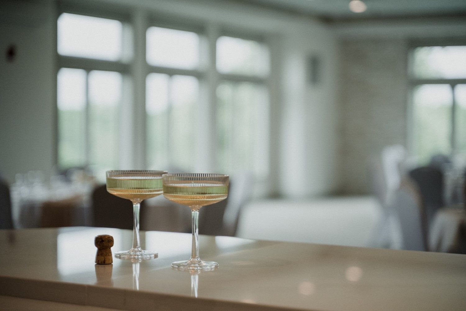 Two champagne coupes on a counter, inside a bright room with windows and chairs in the background.