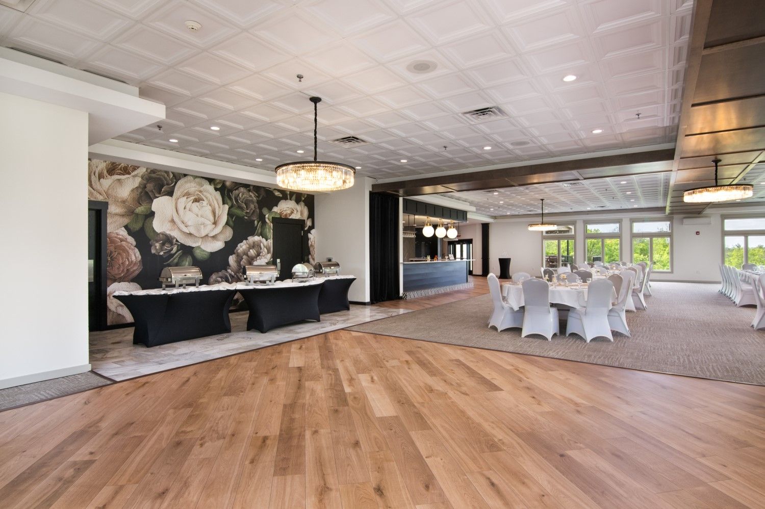 Empty event space with wooden floor, tables set for dinner, floral wall, chandeliers.