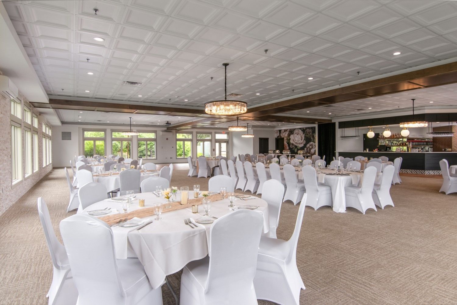An empty wedding reception hall, tables set with white linens, chairs covered, neutral carpet, and an overhead chandelier.