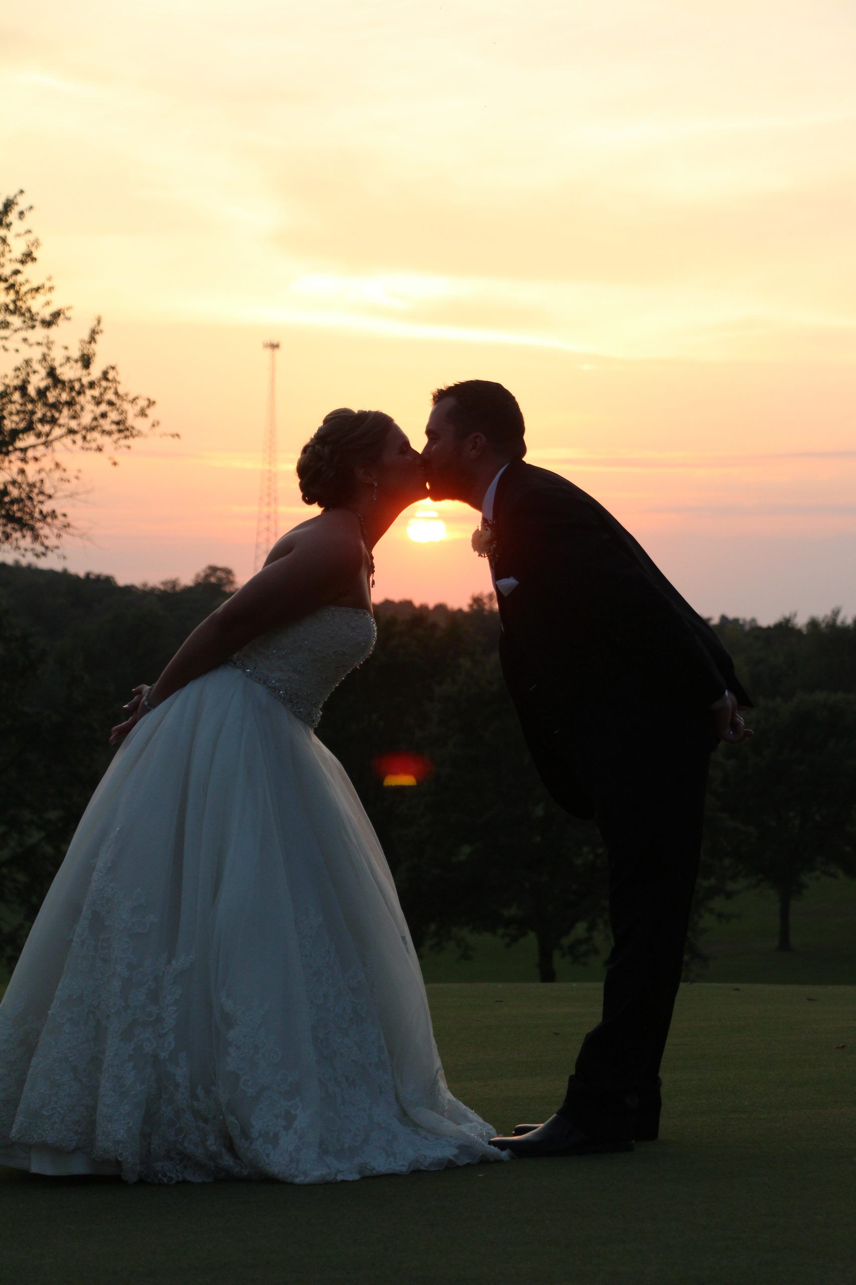 Bride and groom kissing at sunset. Silhouetted on a golf course, with orange sky behind them.