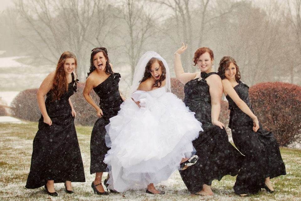 Bride and bridesmaids pose in snow; bride in white gown, bridesmaids in black dresses.