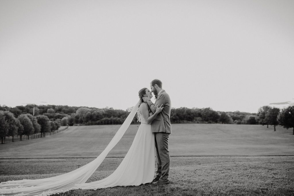 A bride and groom embrace in a field, her long veil trailing behind them. Black and white.