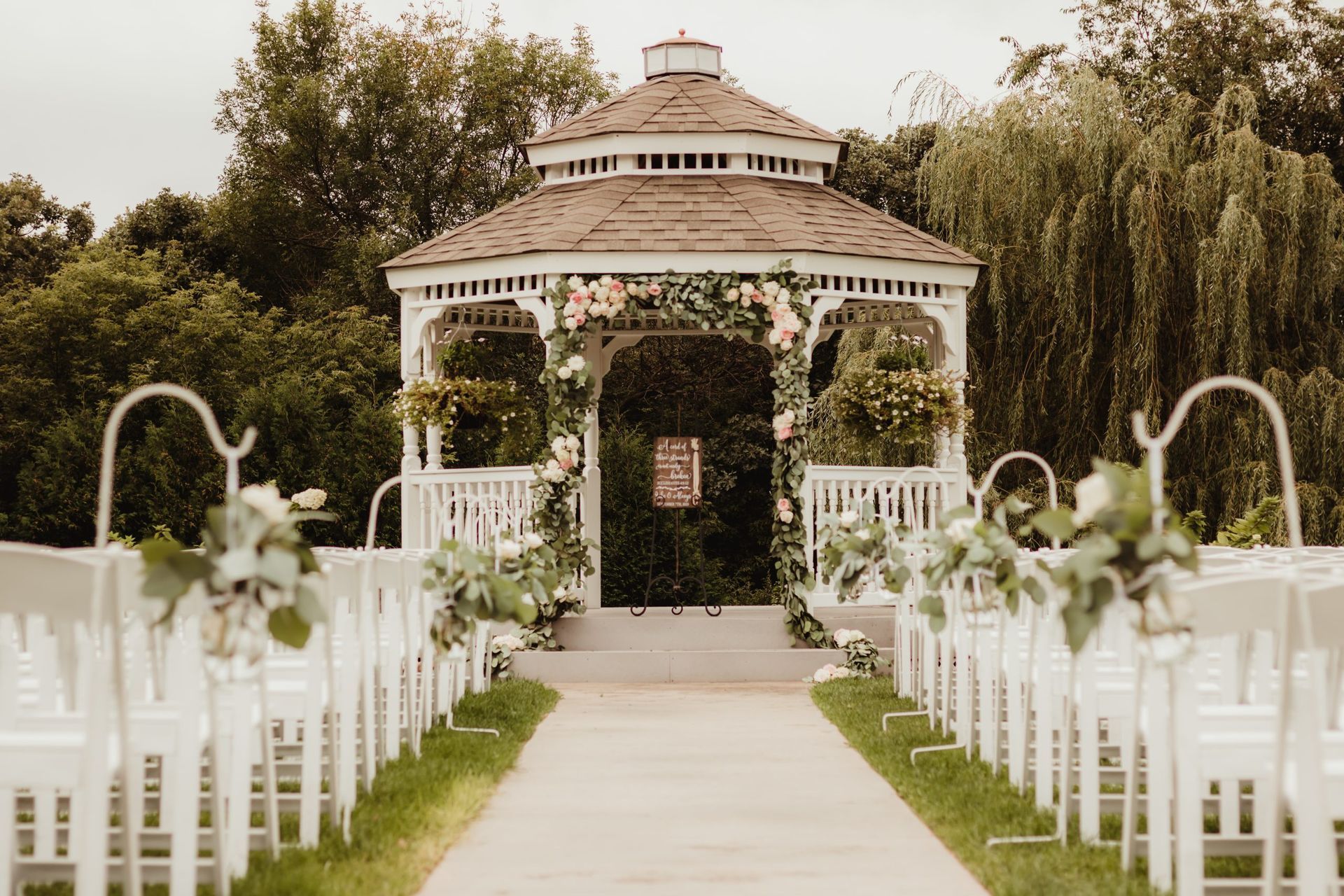 Wedding ceremony setup: White gazebo with floral decorations and rows of white chairs.