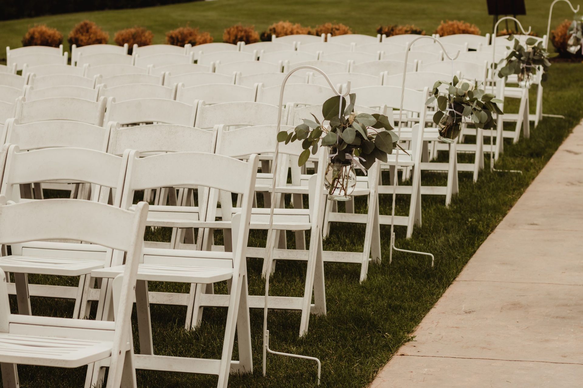 White chairs arranged for an outdoor wedding ceremony on green grass, decorated with floral arrangements.