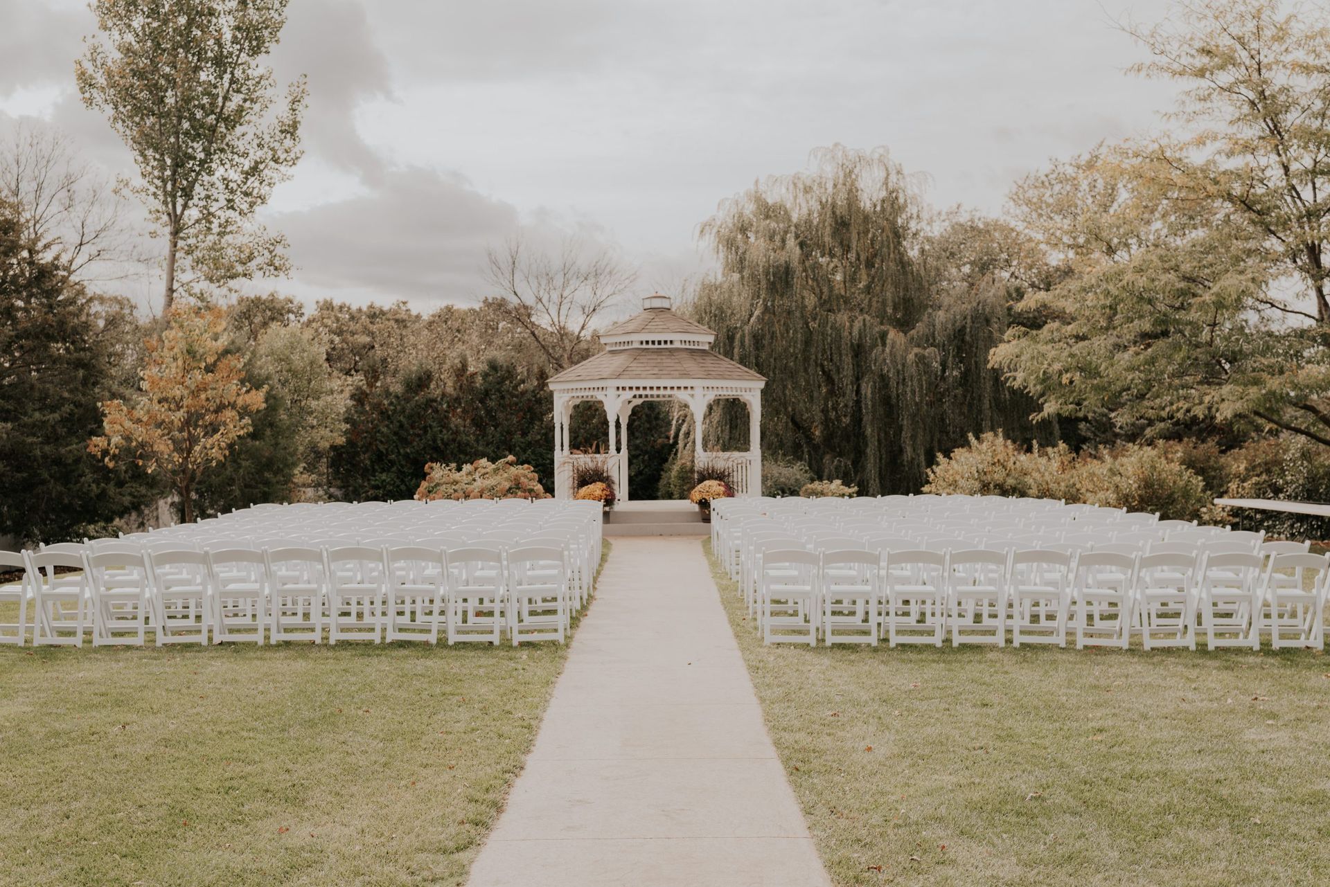 Rows of white chairs face a gazebo on a lawn, pathway leads to the gazebo, trees in background.