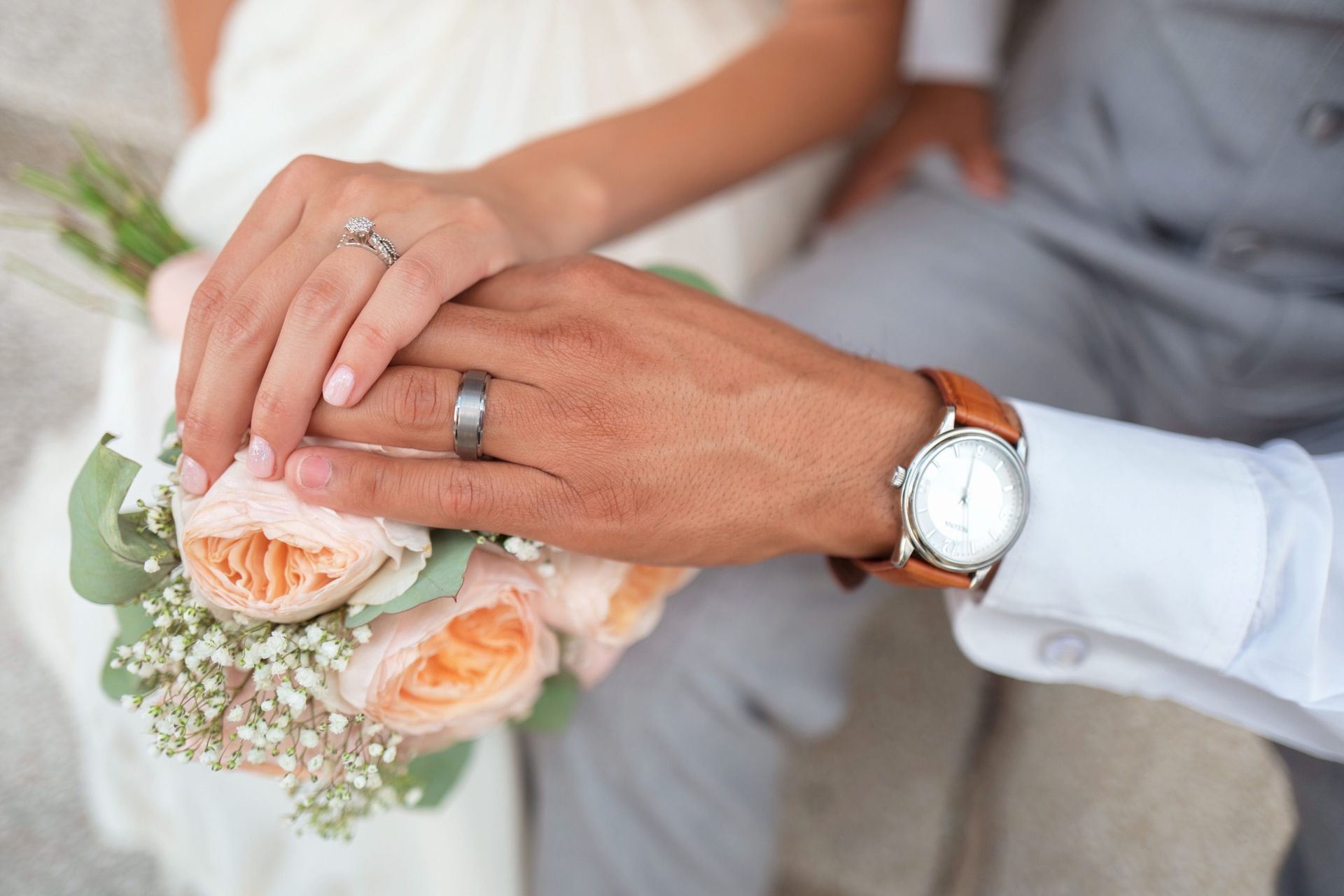 Couple's hands clasped over a bouquet, showing wedding rings and a wristwatch.