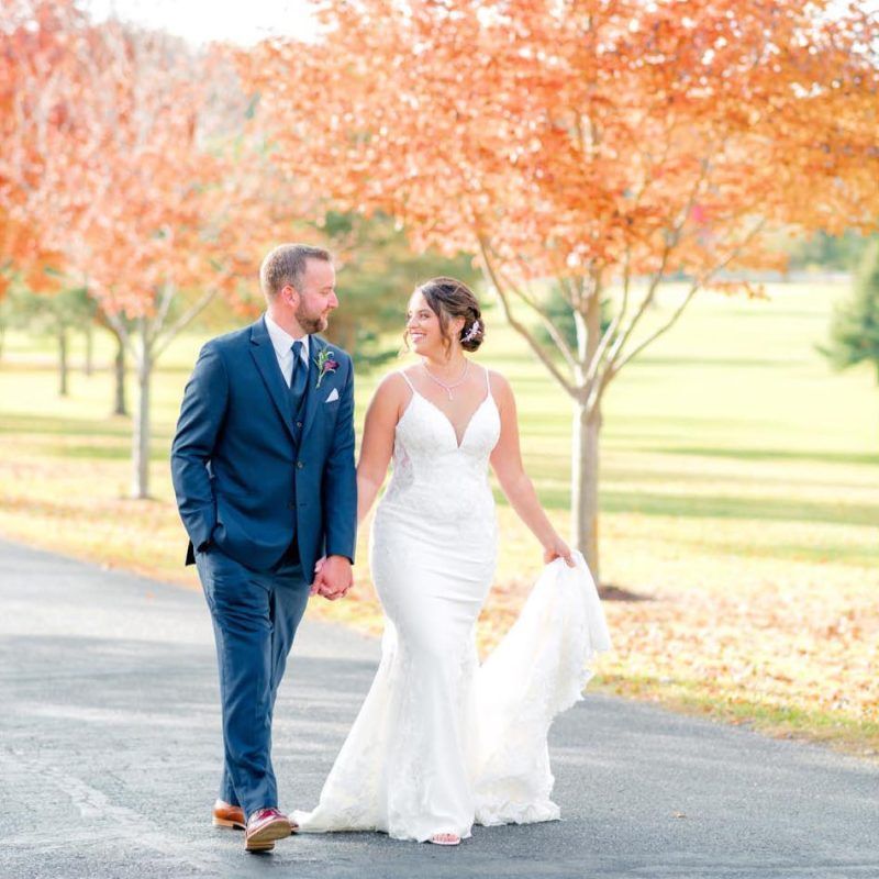 Wedding couple walks down path, holding hands, smiling, with fall trees in background.