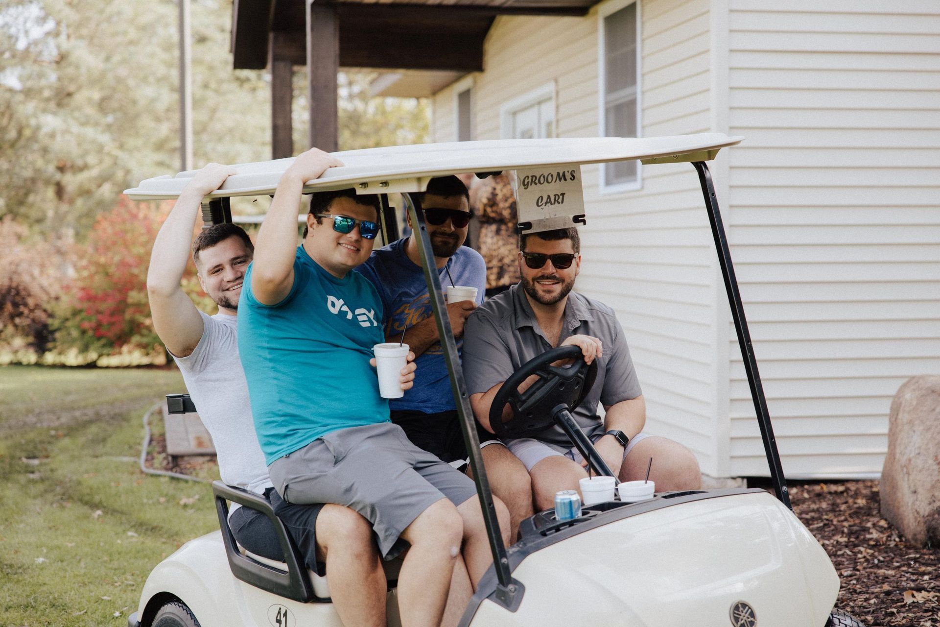 Four men in sunglasses smile in a golf cart parked near a light-colored house.