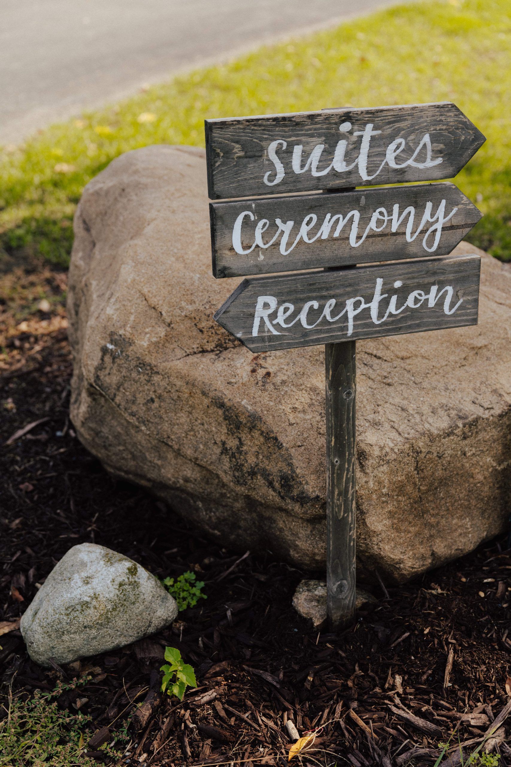 Wooden directional sign with arrows pointing to suites, ceremony, and reception, near a large rock.