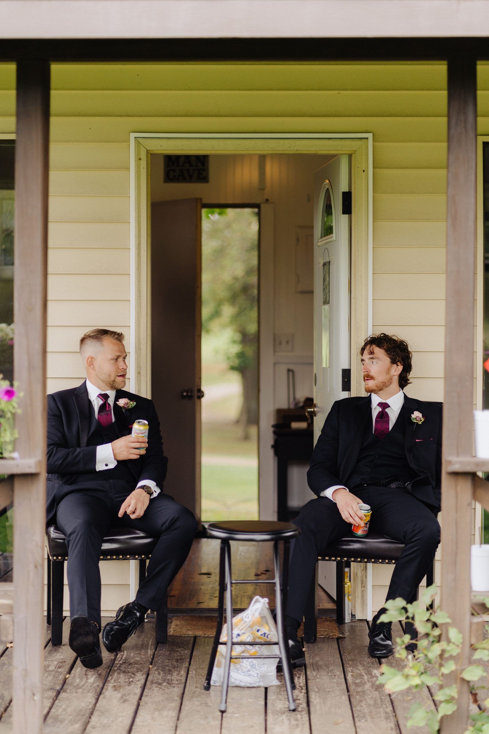 Two men in suits sit on a porch, holding drinks. One looks at the other.