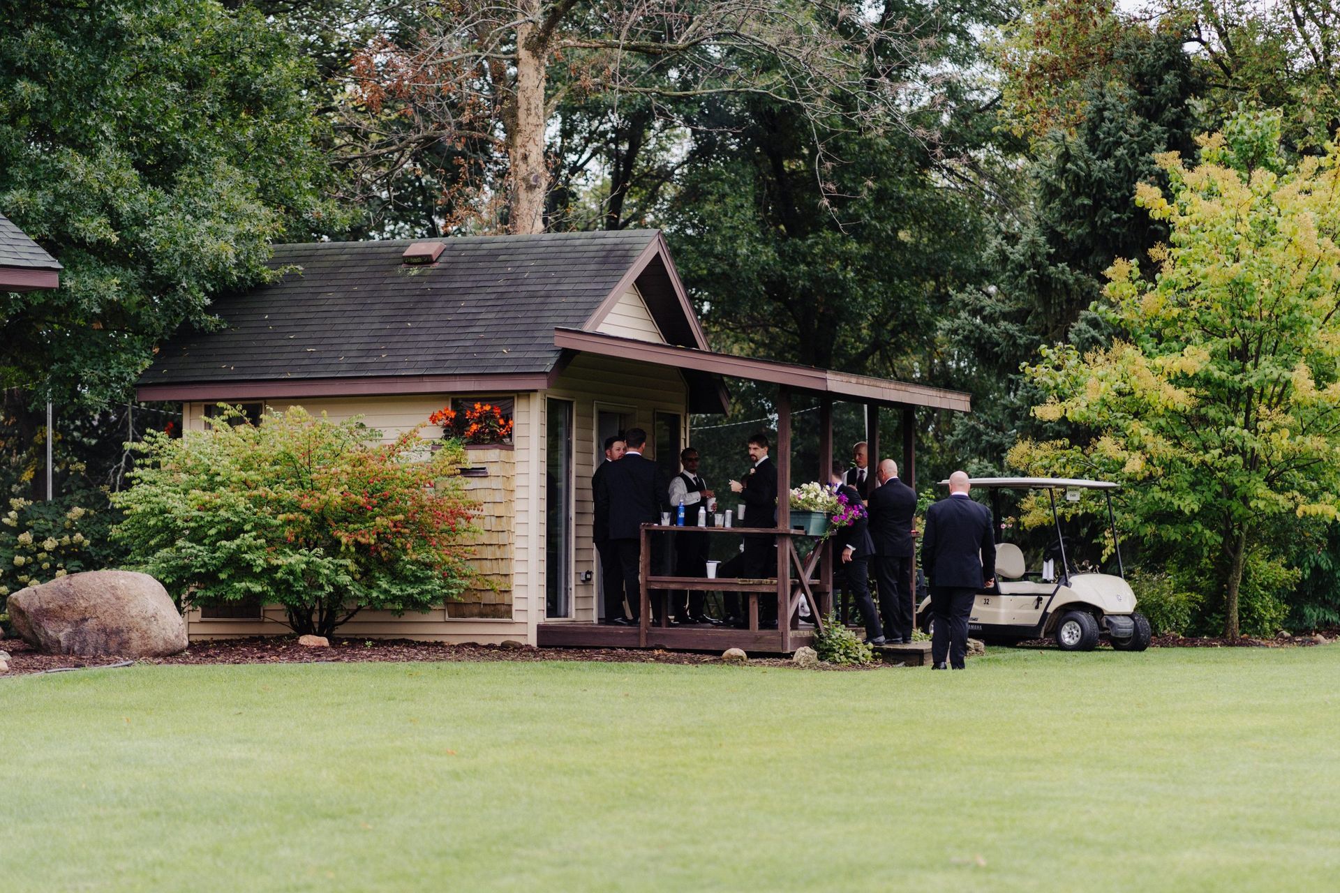 A group of people in dark suits gather near a small wooden cabin with a golf cart on a green lawn.