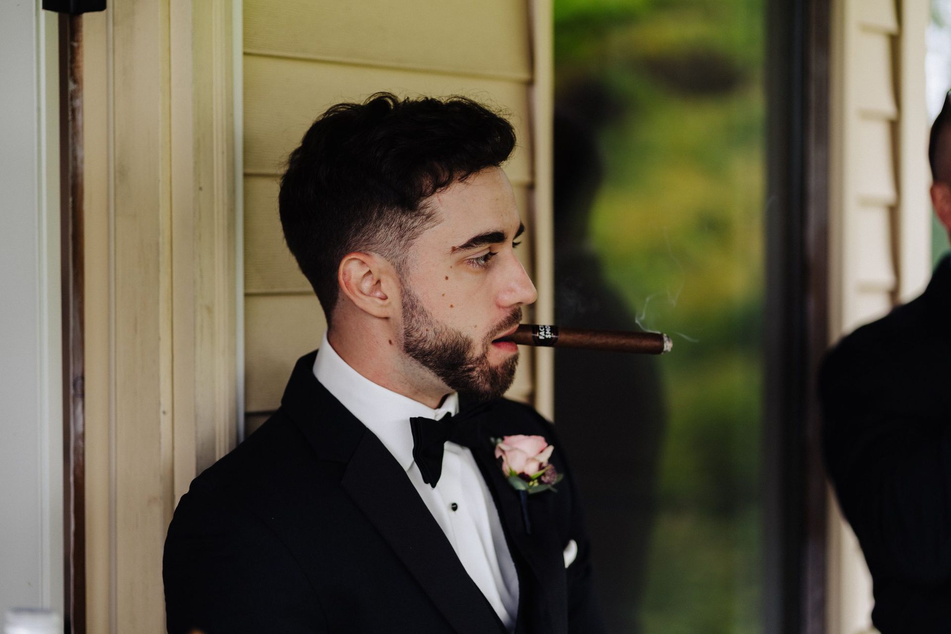 Man in tuxedo smoking cigar, leaning against a building, likely at a wedding.