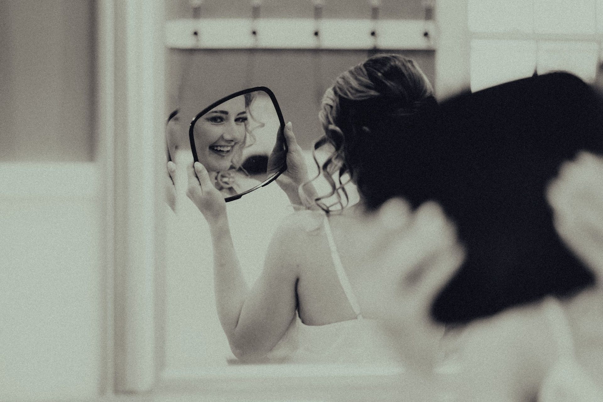 Woman holding a mirror, smiling at her reflection, in a white-walled room.